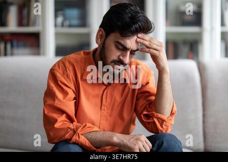 Uomo mediorientale depresso che toccava la testa seduto sul divano di casa Foto Stock