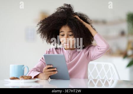 Studentessa nera con afro che indossa una camicia rosa a maniche lunghe che tocca la testa con un tablet seduto a tavola con un piatto di biscotti a casa Foto Stock