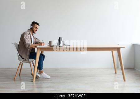 Uomo del Medio Oriente che indossa le cuffie scrivendo appunti mentre lavora su un computer portatile seduto al tavolo in ufficio Foto Stock