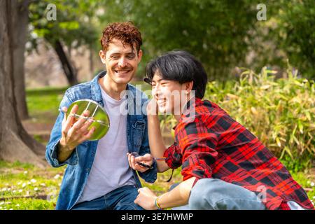 Uomo gay asiatico che guarda il mirrror applicando make up con partner gay caucasico seduto insieme sull'erba nel parco durante il giorno Foto Stock