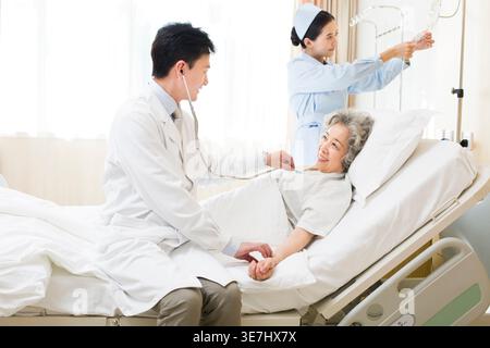 Medico maschile cinese sorridente che utilizza lo stetoscopio per paziente donna cinese anziano sdraiato sul letto vicino infermiera cinese in ospedale Foto Stock
