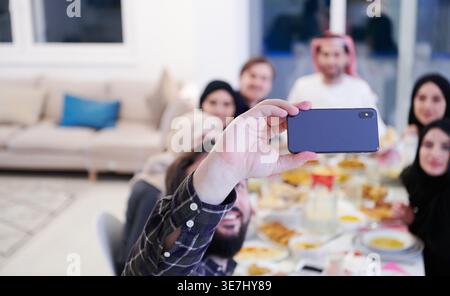 Famiglia musulmana sorridente seduta a tavola a scattare selfie sullo smartphone prima di cenare Iftar durante il mese di festa del Ramadan a casa Foto Stock