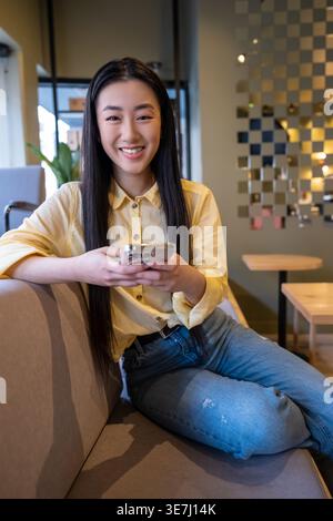 Donna asiatica sorridente che usa lo smartphone mentre siede sul divano in un bar Foto Stock