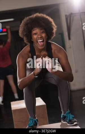 Allegra donna nera con afro e bocca aperta che fa un'esercitazione di salto in scatola alla palestra crossfit Foto Stock