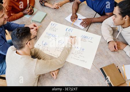 Gruppo di studenti immigrati seduti a tavola che imparano verbi frastagliati durante la lezione di inglese in biblioteca Foto Stock