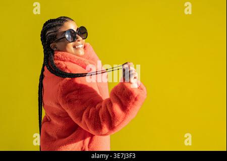 Giovane donna afroamericana con occhiali da sole trecce e cappotto arancione, con sfondo colorato Foto Stock