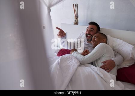 Allegra razza mista e coppia gay caucasica che abbracciano e coccolano mentre sdraiati a letto a casa Foto Stock
