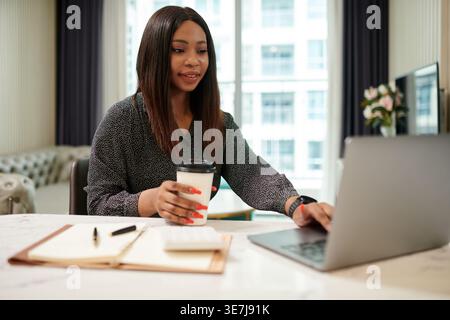 Donna d'affari nera sorridente che tiene in mano il caffè e utilizza un computer portatile mentre si siede al tavolo con un notebook in ufficio Foto Stock