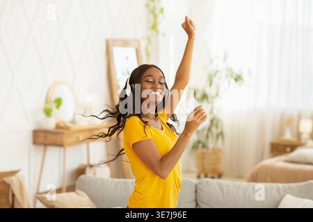 Allegra donna nera con cuffie wireless e smartphone in mano ama ascoltare musica mentre balla a casa Foto Stock