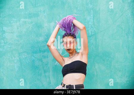 Donna nera con occhiali da sole in piedi con le braccia rialzate che reggono trecce viola contro le pareti turchesi in strada Foto Stock