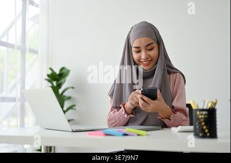 Donna asiatica sorridente in foulard con smLifestylephone seduto vicino al laptop al tavolo in ufficio Foto Stock