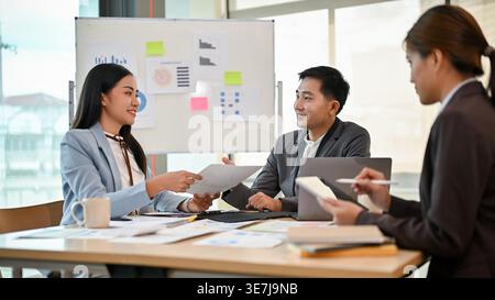 Uomo d'affari asiatico sorridente che lavora insieme a colleghi seduti a tavola e che discute relazioni finanziarie in ufficio Foto Stock