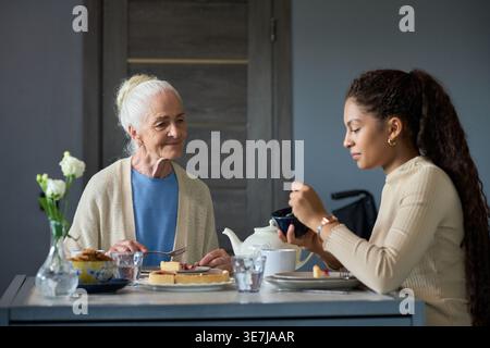 Adolescente nera che tiene la ciotola seduta a tavola mangiando torta con la nonna bianca in cucina a casa Foto Stock