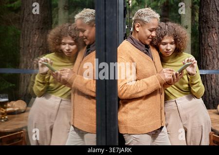 Amiche di corse miste e caucasiche che guardano le foto sullo smartphone mentre sono in piedi e si appoggiano contro la porta di vetro della cabina nella foresta Foto Stock