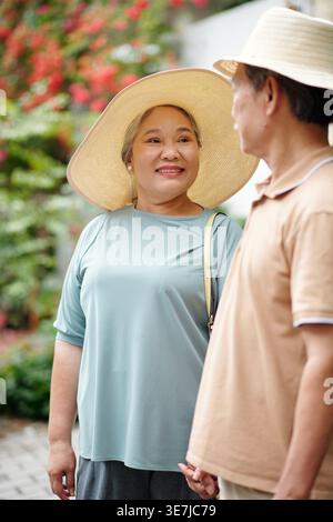 Felice donna asiatica anziana che indossa un cappello di paglia che guarda suo marito mentre si trova in piedi contro il Bush all'aperto Foto Stock