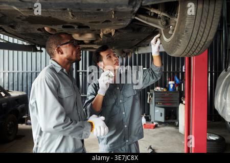 Meccanici maschi neri e asiatici che indossano guanti che riparano sotto il telaio dell'auto mentre si trova in un'officina di riparazione auto Foto Stock