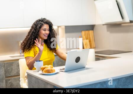 Donna ispanica con capelli ricci che partecipa a una riunione online utilizzando un computer portatile mentre fa colazione al bancone della cucina a casa Foto Stock