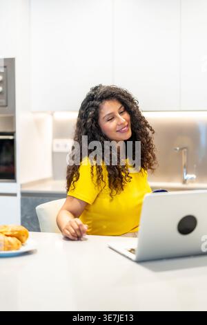 Donna ispanica con capelli ricci che partecipa a una riunione online utilizzando un computer portatile mentre fa colazione al bancone della cucina a casa Foto Stock