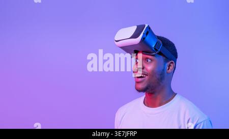 Uomo nero che indossa una camicia bianca e un visore per la realtà virtuale che guarda lontano mentre sta in piedi sullo sfondo viola al neon in studio Foto Stock
