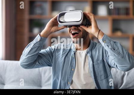 Uomo nero che indossa occhiali VR e goditi un'esperienza di realtà virtuale seduto sul divano di casa Foto Stock