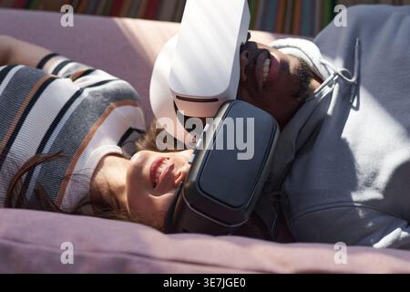 Coppia nera e mista che indossa il visore VR sul divano in salotto di casa Foto Stock