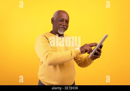 Uomo Nero di mezza età che indossa un maglione giallo che regge un tablet mentre si trova su sfondo giallo in studio Foto Stock