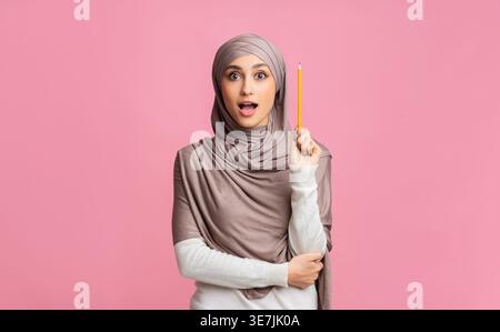 Donna araba che indossa un hijab che tiene in piedi una matita con le braccia incrociate su sfondo rosa Foto Stock