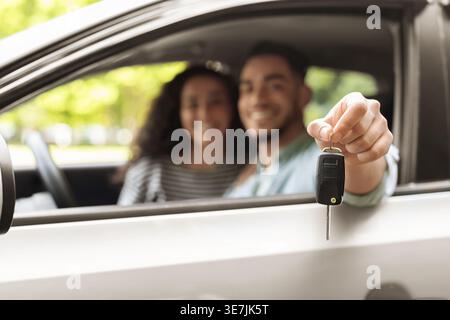 Sorridente uomo mediorientale che tiene in mano la chiave con telecomando mentre è seduto in auto con la sua ragazza durante il giorno di sole Foto Stock