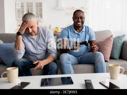 Uomo nero e amico di mezza età caucasico maschio che si diverte a giocare con i controller mentre si siede sul divano nel soggiorno di casa Foto Stock