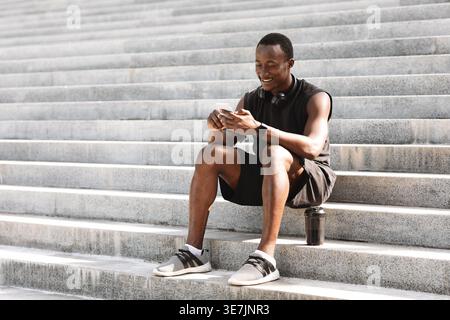 Uomo nero che usa lo smartphone seduto sulle scale all'aperto nel parco nelle giornate di sole Foto Stock