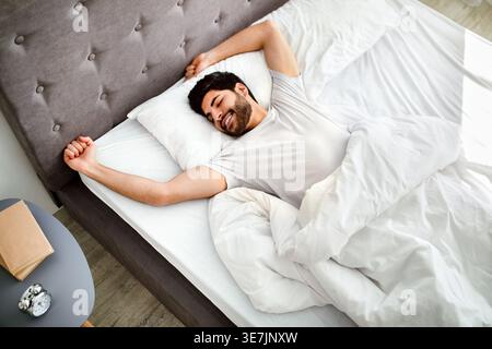Uomo arabo con barba che indossa una camicia bianca che allunga le braccia con gli occhi chiusi mentre si sdraia sul letto in camera da letto di casa Foto Stock
