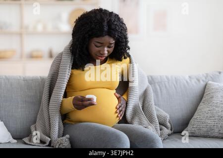 Donna incinta di colore con capelli ricci che indossano maniche lunghe gialle avvolta in coperta che le toccano la pancia soffre di dolore al travaglio seduta sul divano... Foto Stock