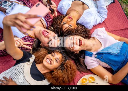 Allegro e variegato gruppo di amiche che scattano selfie con lo smartphone mentre si sdraia sulla coperta nel parco Foto Stock