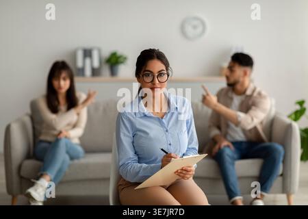 Psicologa donna mediorientale che indossa occhiali da vista e scrive sugli appunti mentre si siede contro coppie di pazienti che discutono in clinica Foto Stock