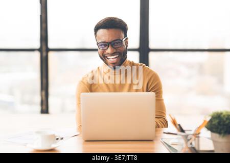 Allegro uomo nero che indossa occhiali da vista seduto a un tavolo con un computer portatile in ufficio Foto Stock
