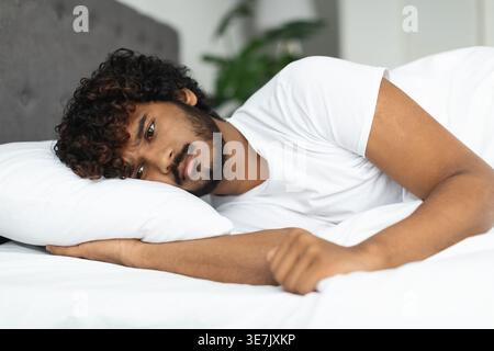 Un triste indiano barbuto con capelli ricci sdraiato sul letto in camera da letto di casa Foto Stock