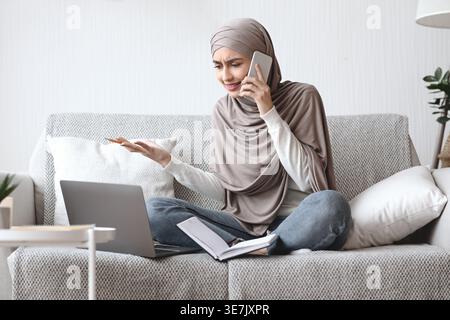 Una donna musulmana araba delusa in un hijab che gestiva la mano mentre faceva una telefonata seduta con le gambe incrociate sul divano con un portatile e un notebook a casa Foto Stock