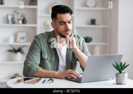 Uomo arabo che tocca il mento guardando lo schermo del notebook seduto a un tavolo a casa Foto Stock