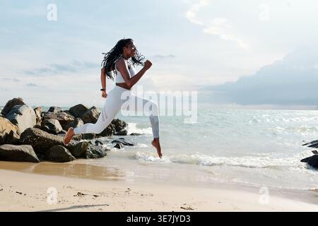 Donna nera con dreadlock che indossa un outfit fitness che corre in riva al mare durante la giornata di sole Foto Stock