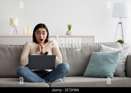 Una donna araba scioccata con le mani strette che guarda lo schermo di un laptop seduto con le gambe incrociate sul divano nel soggiorno di casa Foto Stock