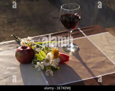 Bicchiere di vino rosso con mela e fiori selvatici su una tovaglia a scacchi, illuminato da una calda luce dorata in un ambiente tranquillo all'aperto. 2026 Foto Stock