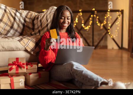 Adolescente nera con laptop che tiene la carta di credito seduta con gambe incrociate allungate sul pavimento nel soggiorno di casa Foto Stock