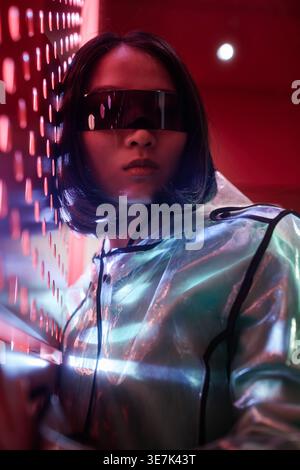 Donna asiatica che indossa visiera futuristica e giacca iridescente in studio con luci al neon Foto Stock
