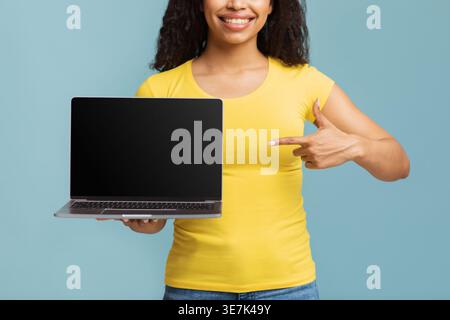 Donna nera sorridente che indossa una camicia gialla che mostra e indica il laptop in piedi su sfondo blu in studio Foto Stock