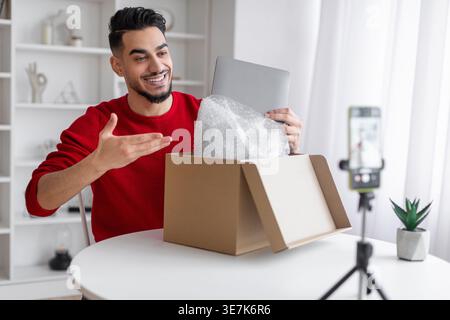 Influencer maschile arabo che registra contenuti video utilizzando lo smartphone che disimballano il pacco con il laptop seduto al tavolo di casa Foto Stock