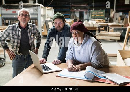 Donna nera in piedi con carpentieri caucasici che utilizza un computer portatile in un negozio di lavorazione del legno Foto Stock