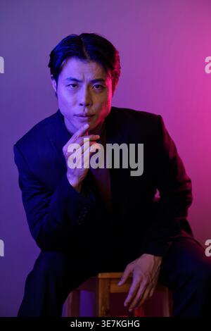 Uomo asiatico che si appoggia al mento a mano appoggiato in avanti seduto su una sedia di legno in studio con luci al neon rosa e viola Foto Stock