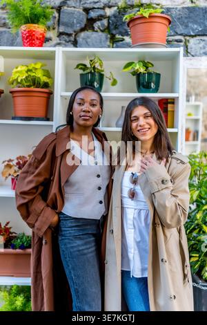Amiche nere e caucasiche che indossano trench e scelgono piante sugli scaffali mentre si trovano in città durante il giorno Foto Stock