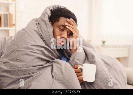 Uomo nero malato avvolto in una coperta che regge una tazza di caffè mentre seduto a occhi chiusi sul divano di casa Foto Stock