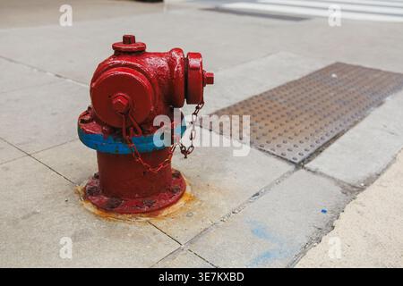 Un idrante antincendio si trova sul marciapiede vicino all'angolo di una strada. E' dipinto di rosso con accenti blu. L'idrante è dotato di una catena metallica collegata ed è impostato Foto Stock